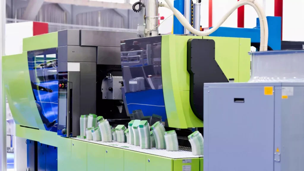 A modern green and black injection molding machine in a production facility, actively processing plastic containers on a conveyor belt