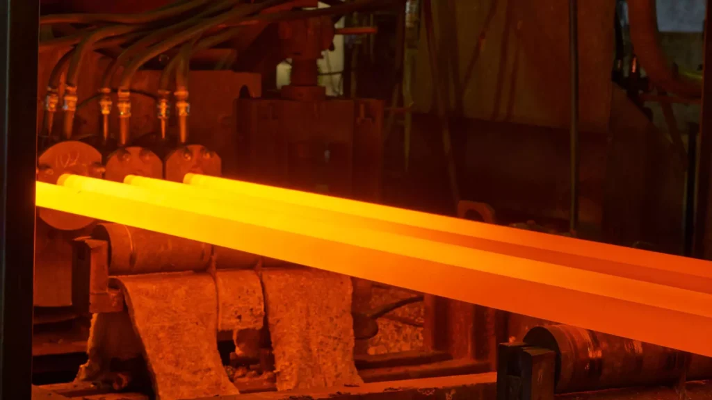 Glowing hot steel bars moving along rollers in a forging process, with industrial machinery shaping the metal under intense heat.