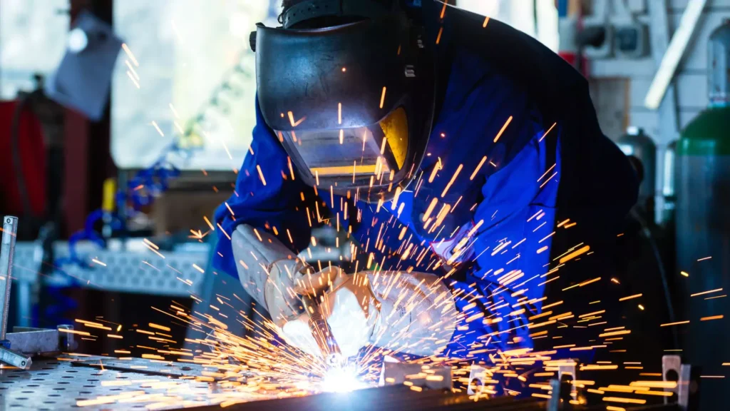 A welder wearing a protective mask and gloves, working with metal while bright sparks fly in a factory setting.