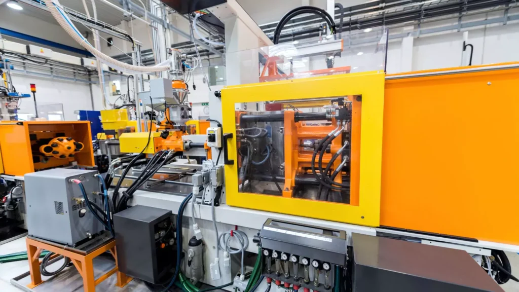 An industrial orange and yellow injection molding machine in a factory setting, showcasing its mechanical components and hydraulic systems