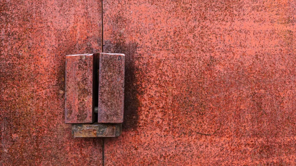 A close-up of a heavily rusted metal door with a corroded locking mechanism, showing signs of weathering and oxidation.