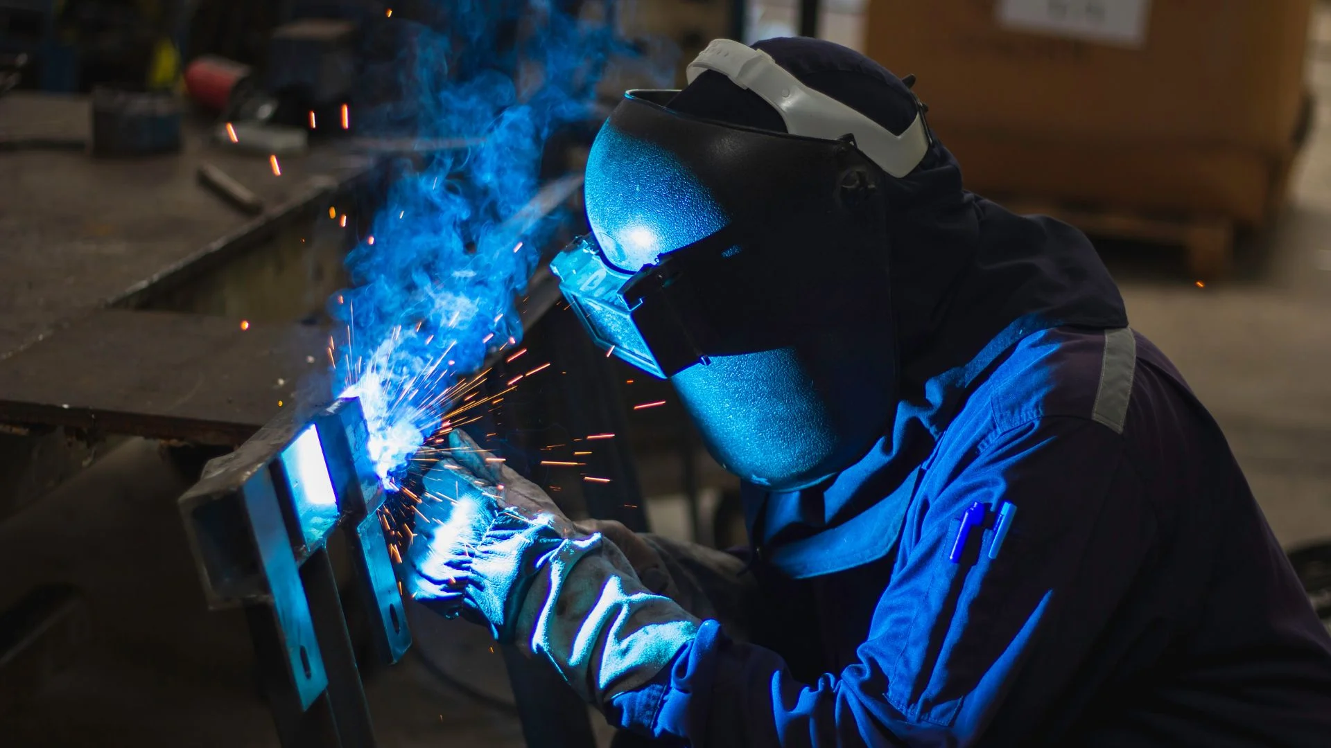 A welder in full protective gear working on a metal structure, with blue welding arcs, smoke, and sparks illuminating the dark workshop environment.