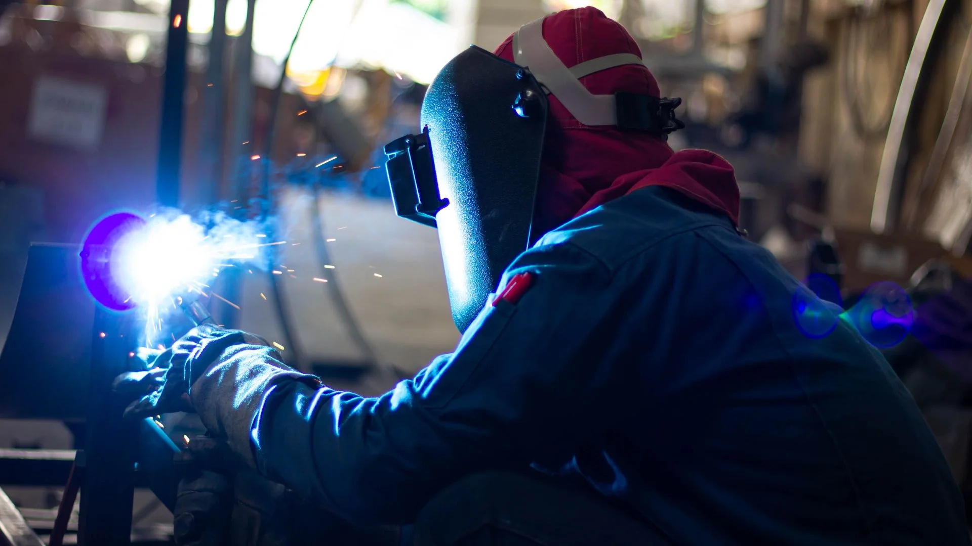 A welder wearing protective gear and a red head covering, performing metal fabrication welding with bright blue sparks and smoke in an industrial workshop.