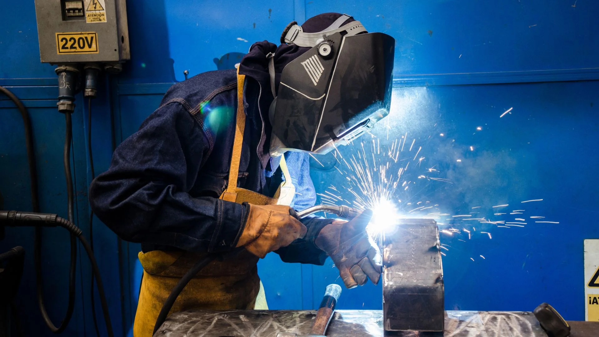 A professional welder wearing protective gear and a welding helmet, working on a metal structure with bright sparks flying in a workshop.