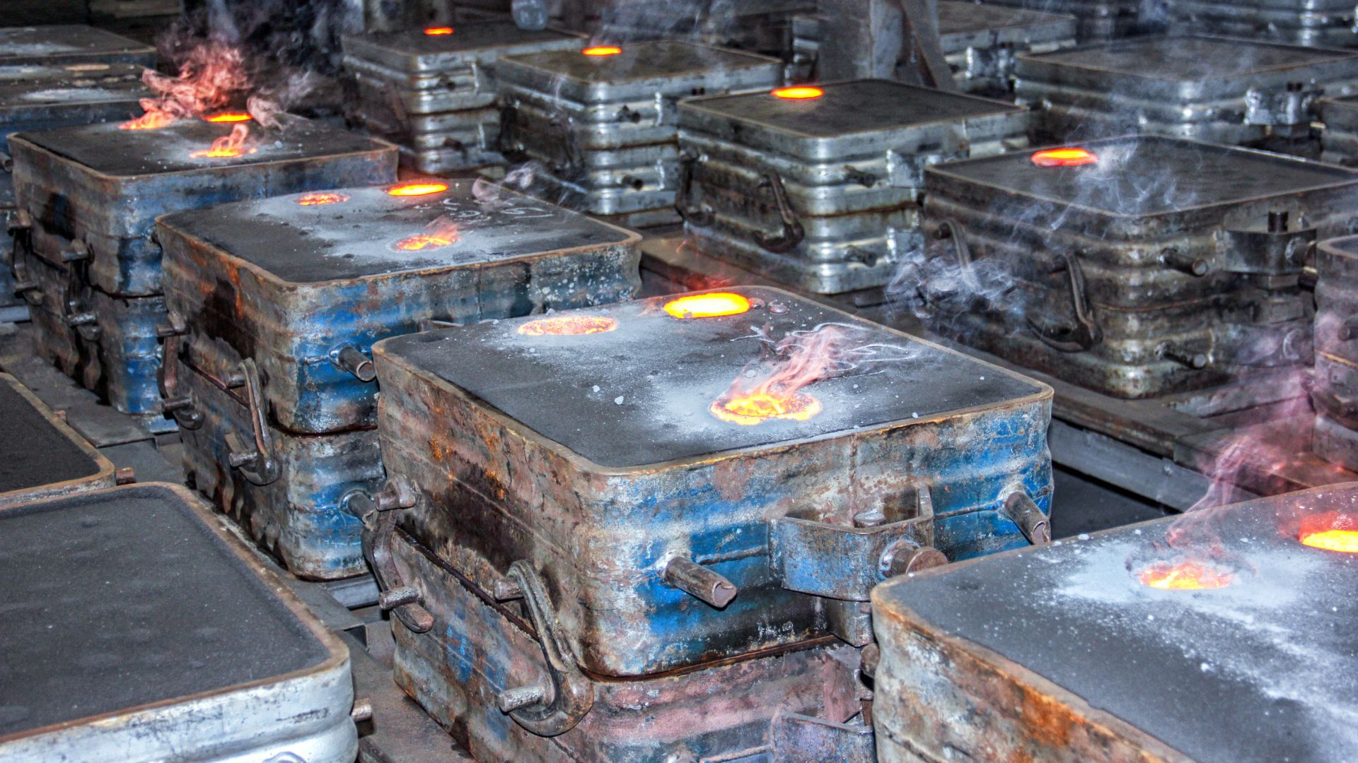 A foundry scene showing multiple sand casting molds with molten metal being poured, glowing with heat and emitting smoke during the solidification process.